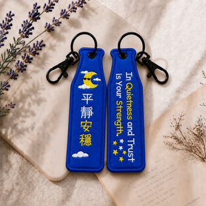 平靜藍色瓶安同行刺繡吊飾雙面展示,一面為中文「平靜安穩」字樣,另一面為英文 In Quietness and Trust is Your Strength. 字樣,搭配月亮、雲朵與星星刺繡及黑色掛鉤。