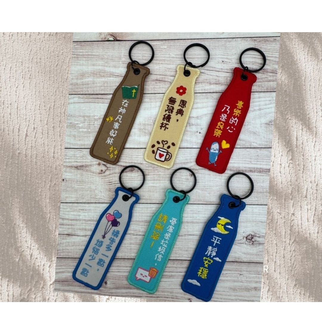 瓶安同行刺繡吊飾系列中文面展示，多款不同色系吊飾排列於木紋背景上，呈現喜樂、平靜安穩、在神凡事都能、恩典無限續杯等中文祝福文字。
