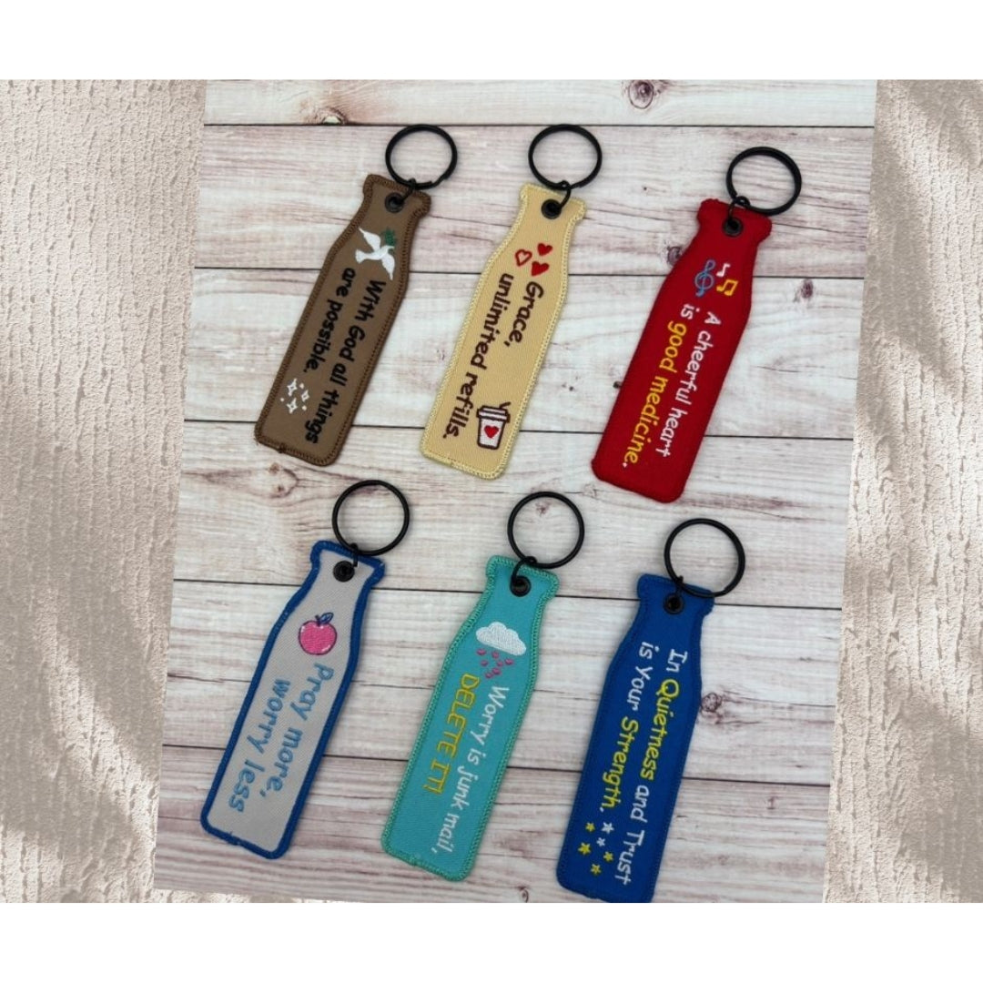 瓶安同行刺繡吊飾系列英文面展示，多款不同色系吊飾排列於木紋背景上，呈現 A cheerful heart is good medicine、Pray more worry less、With God all things are possible 等英文祝福文字。