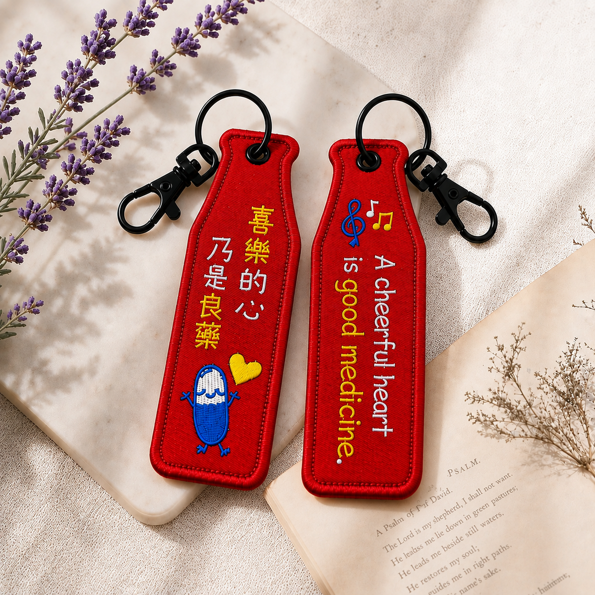 喜樂紅色瓶安同行刺繡吊飾雙面展示，一面為「喜樂的心乃是良藥」中文祝福文字，另一面為 A cheerful heart is good medicine 英文文字，搭配音符、愛心與黑色掛鉤。