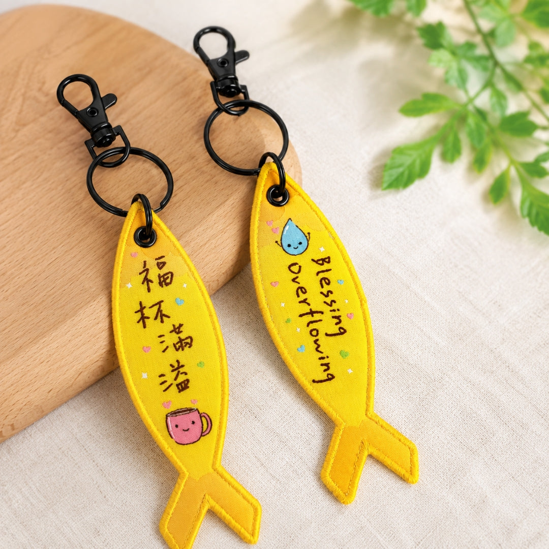 福杯滿溢黃色得人如魚吊飾雙面展示，魚形彩織吊飾分別呈現中文「福杯滿溢」與英文 Blessing Overflowing 字樣，搭配杯子、水滴插畫及黑色掛鉤。