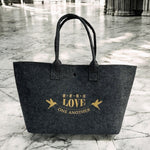 黑色毛氈提袋正面展示，袋身印有金色「LOVE ONE ANOTHER」字樣與彼此相愛圖案，置於灰白大理石地面背景前。