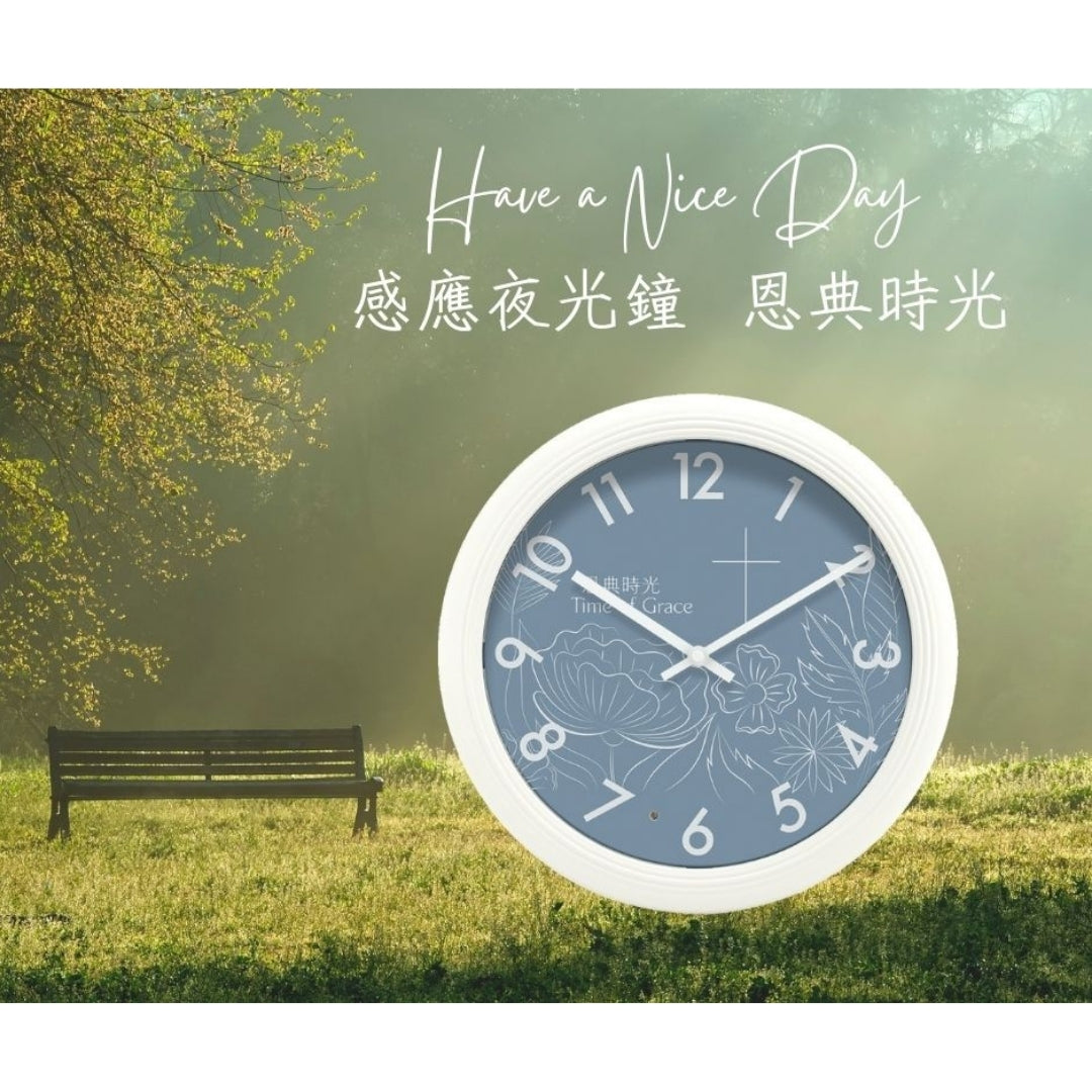 恩典時光感應夜光鐘日間情境圖，白框藍面掛鐘置於綠意草地背景前，呈現日間外觀與清新氛圍。