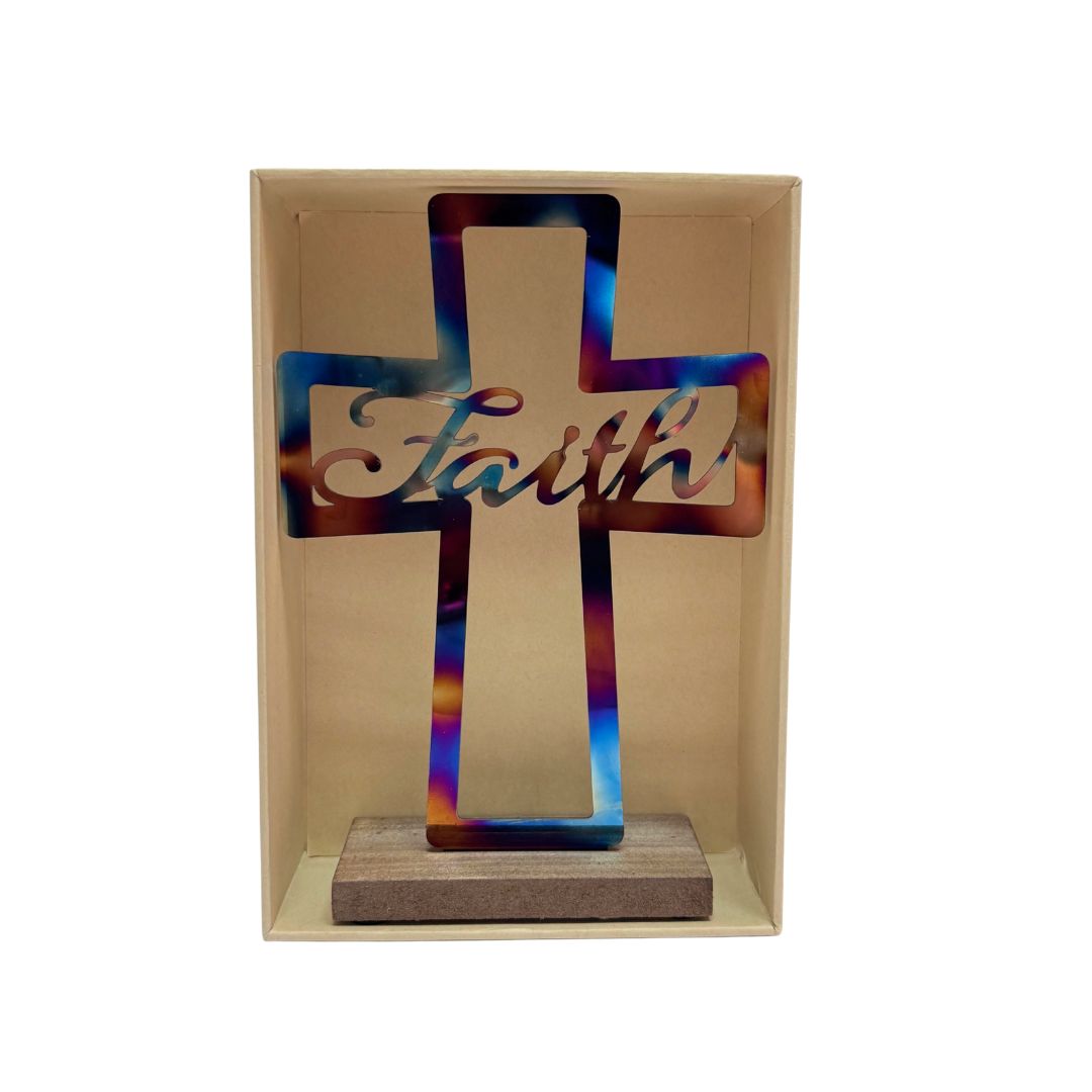 A metal cutout cross with the word 'Faith' in script, displayed inside a clear-front beige shadow box on a wooden base. The cross has an iridescent, rainbow-like reflective finish.