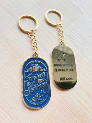 格子外面《出入平安》城市款鑰匙圈正反面展示，正面為藍底金色城市建築及英文 Protects You on Your Journey，背面刻有詩篇121篇8節中文經文。